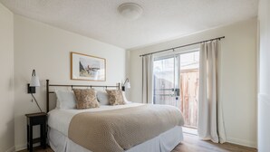 A bedroom with a large bed, a nightstand, a lamp, a framed picture on the wall, and a sliding glass door leading to an outdoor area. - Steps to the Sand + Patio Lounging | Sunset Water Views | Bay View 3 (San Diego)