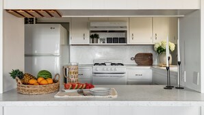 A kitchen with a refrigerator, stove, and cabinets. There is a fruit basket with oranges and a watermelon, and a glass pitcher on the counter. - Steps to the Sand + Patio Lounging | Sunset Water Views | Bay View 3 (San Diego)