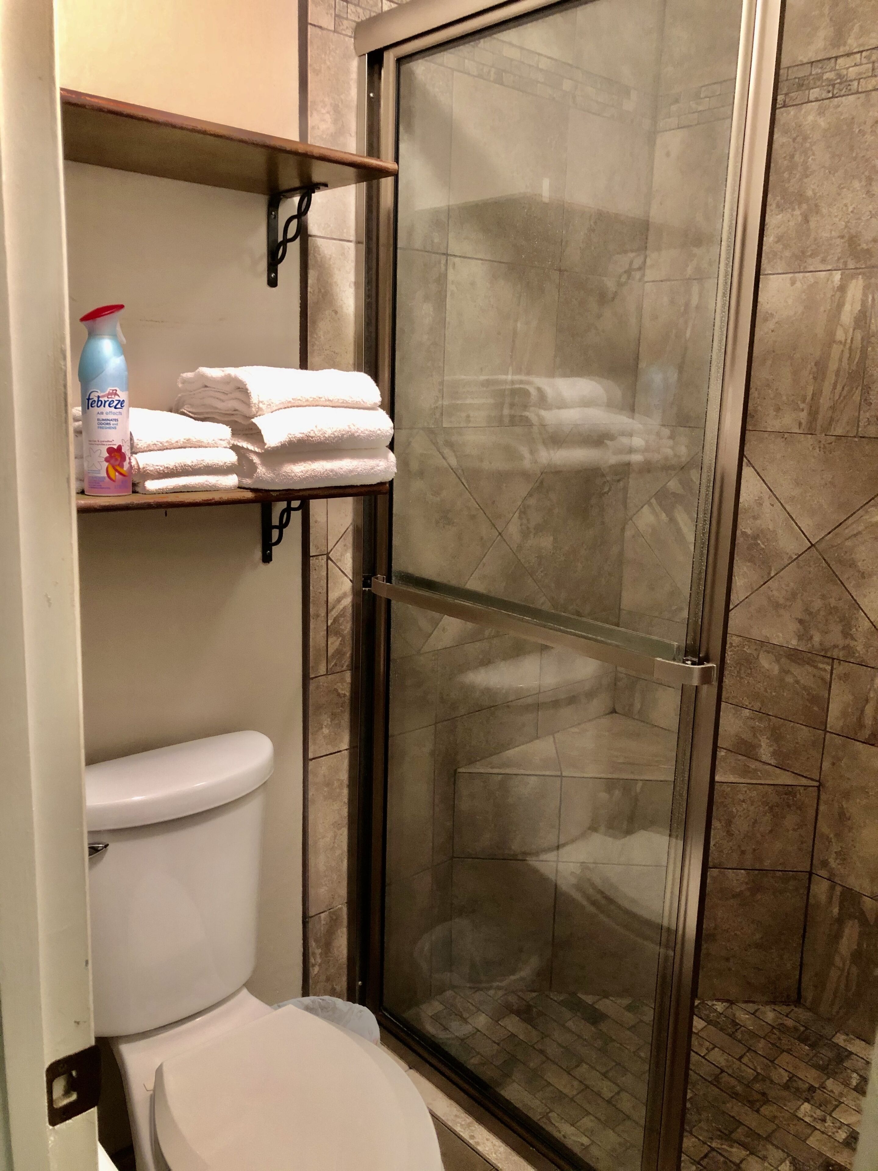 A bathroom with a toilet, a glass-enclosed shower, and a shelf with folded towels and a bottle of hand soap.