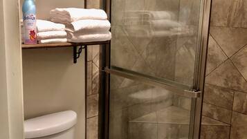 A bathroom with a toilet, a glass-enclosed shower, and a shelf with folded towels and a bottle of hand soap.