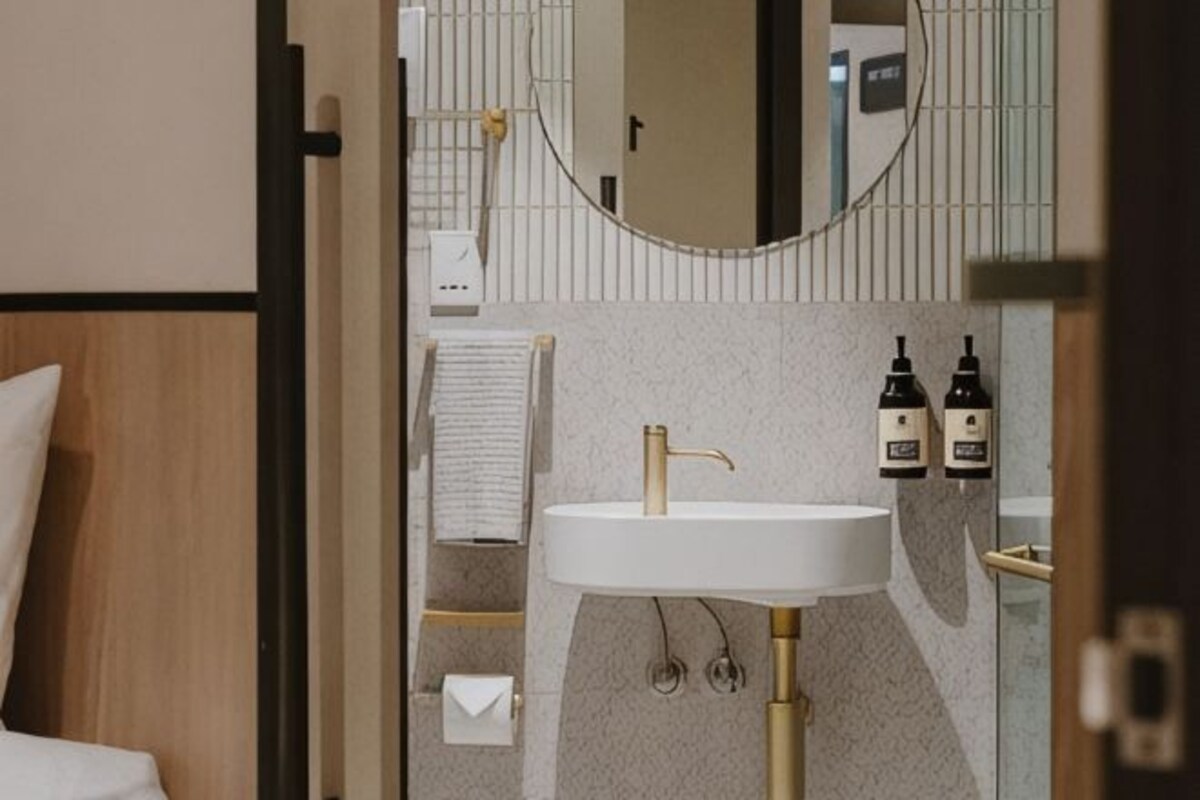 A modern bathroom with a white sink, gold faucet, and a mirror above it. There are two bottles of soap on the wall.