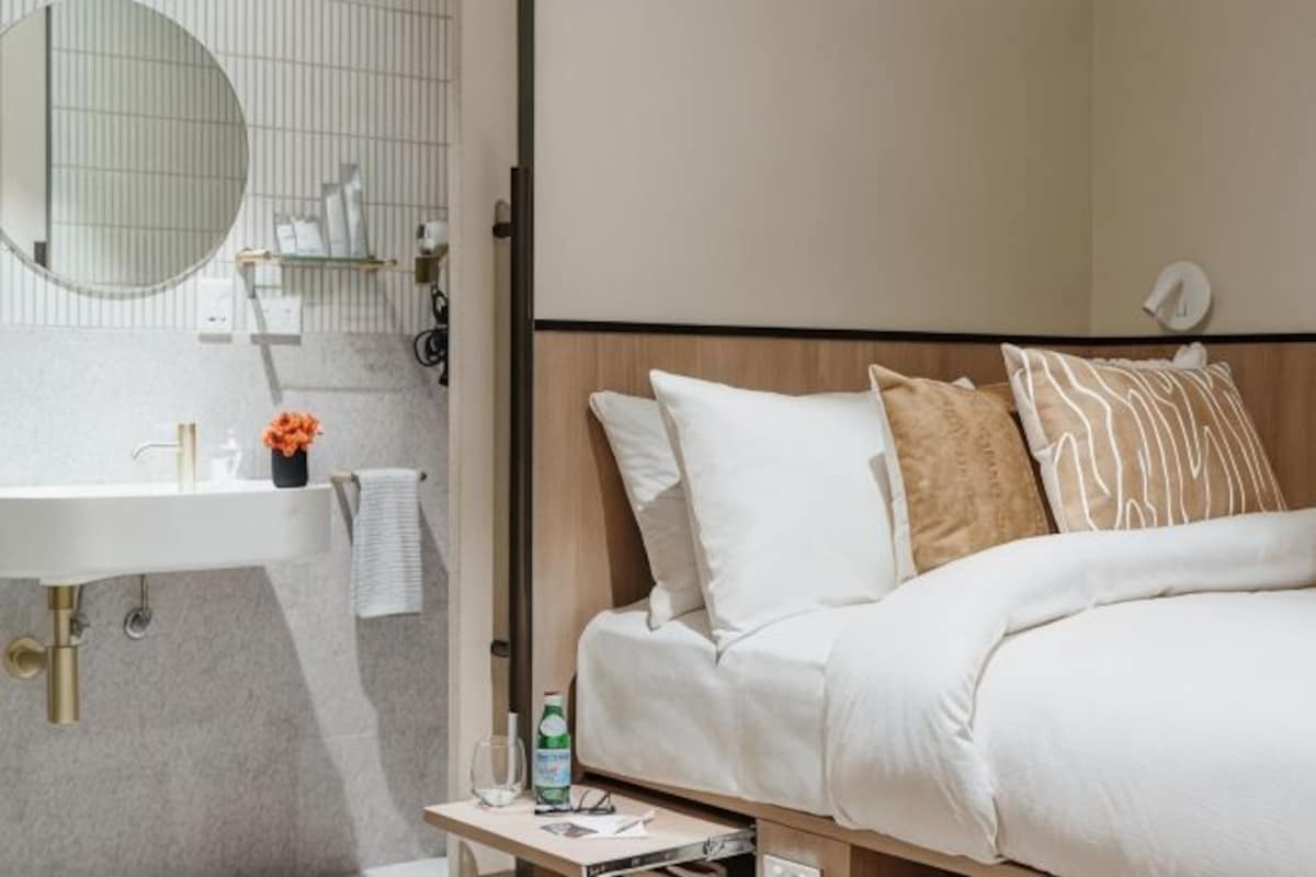 A hotel room with a bed, a bedside table, a mirror, a sink, and a towel rack.