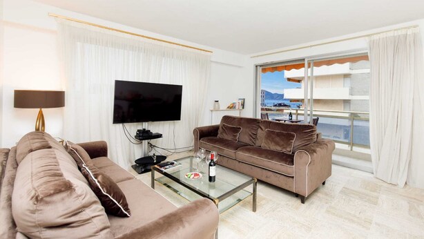A modern living room with a brown sofa, a glass coffee table with a bottle and glasses, a flat-screen TV, and a large window with a view of buildings and mountains. - Appartement Standing, 90m² Climatisé à 5 mns à Pied de la Croisette et Plages (Cannes)