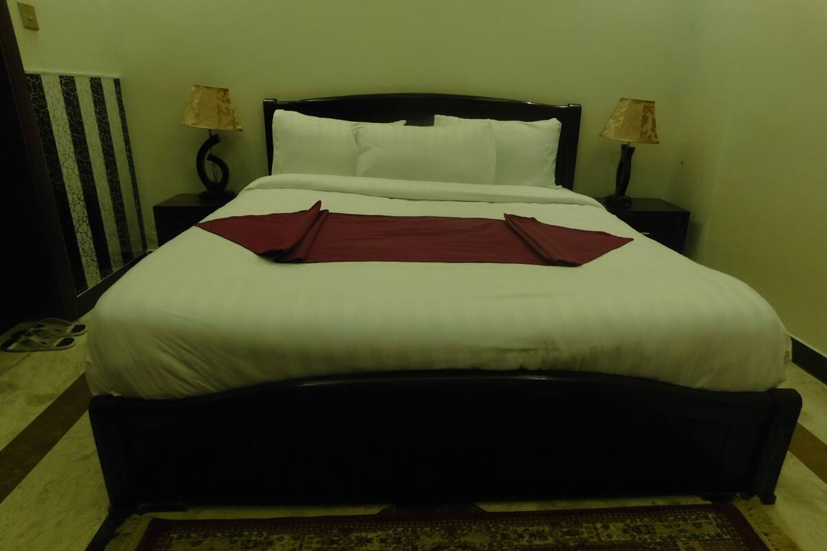 A neatly made bed with white linens and a red throw, a black headboard, two bedside lamps, and a patterned rug on the floor.