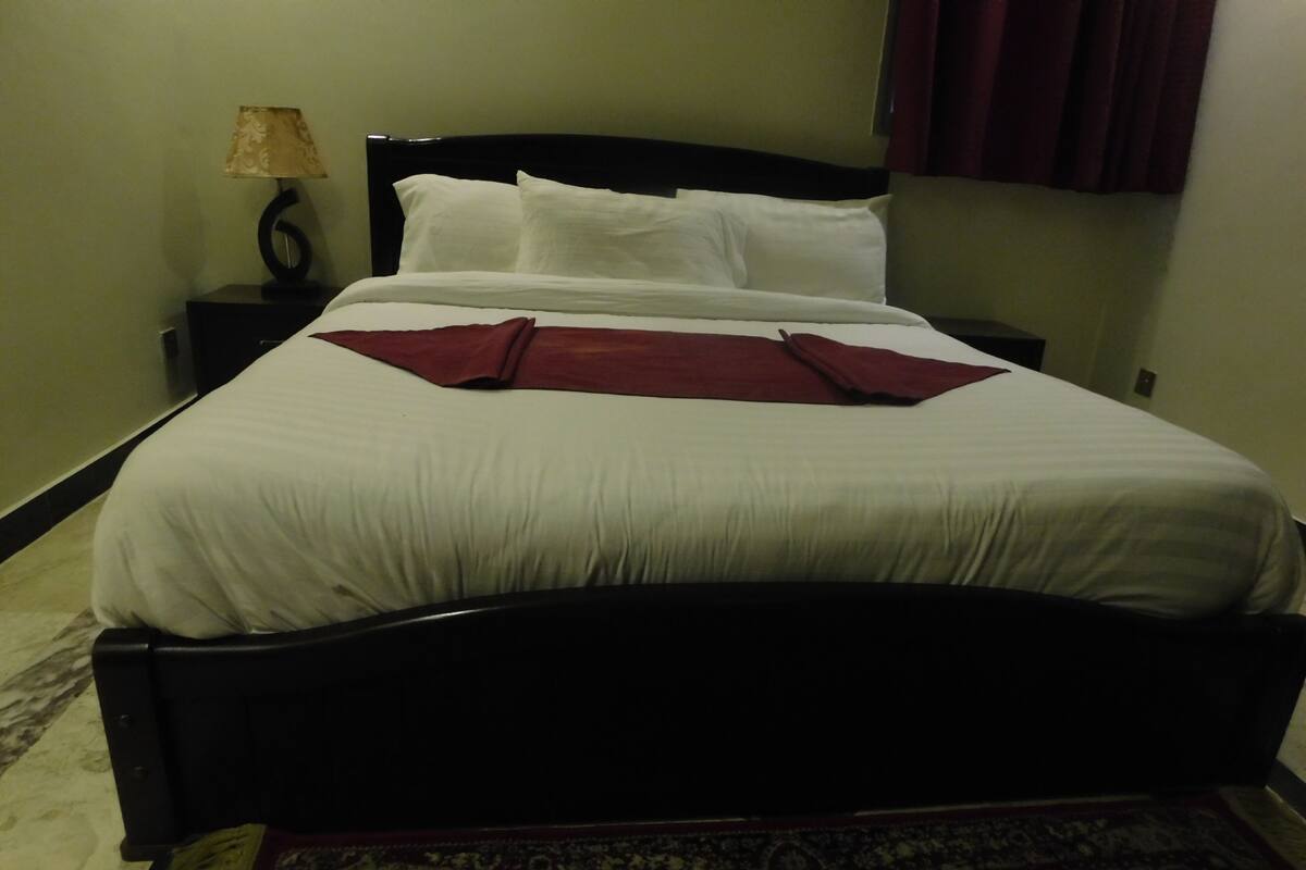 A neatly made bed with white bedding and a red throw blanket, a dark wooden headboard, a small bedside table with a lamp, and a patterned rug on the floor.