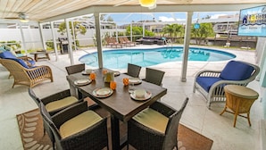 A covered outdoor area with a pool, dining table set with plates and glasses, wicker furniture, and a sign for Roelens Vacations. - Prestigious location, Gulf Access, Heated Pool - Villa Pineapple Oasis - Roelens Vacations (Cape Coral)