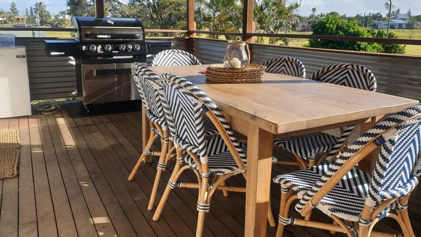 Un patio exterior con mesa de madera, sillas de mimbre, una parrilla y vistas a la vegetación circundante.
