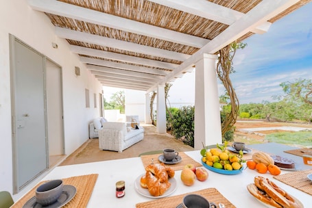 A covered outdoor dining area with a table set for breakfast, including croissants, fruits, and pastries.