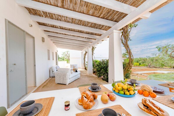 A covered outdoor dining area with a table set for breakfast, including croissants, fruits, and pastries.
