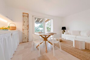 A modern kitchen with white cabinetry, a dining table, and chairs. There is a white sofa and a lamp in the living area.