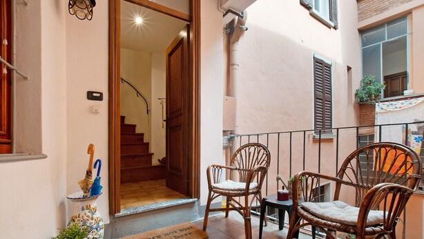 Un balcón con sillones de mimbre, una mesita, un florero decorativo y una puerta que da a un espacio interior.
