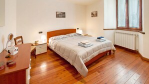 Un dormitorio con cama de madera, mesitas de noche, un escritorio con silla, una ventana con cortinas y dos cuadros en la pared.