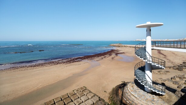 A spiral staircase leading to a beach with a clear view of the ocean and a pier in the background.