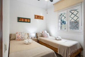 Une chambre avec deux lits simples, une petite table de chevet avec une lampe, une fenêtre ornée d’un motif en treillis et une œuvre d’art accrochée au mur.