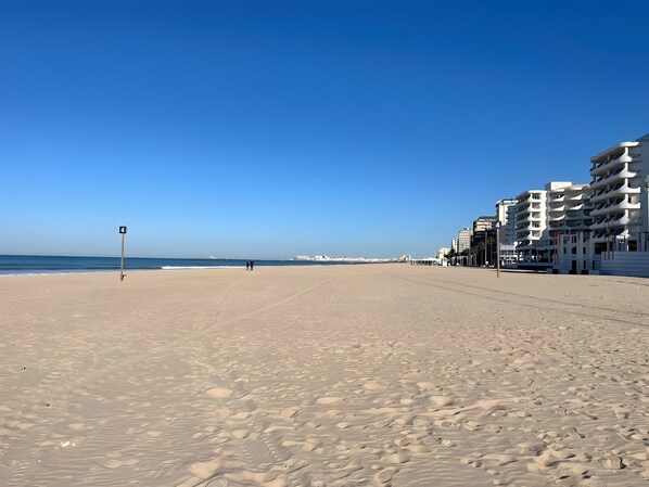 Une vaste plage de sable fin, un ciel bleu dégagé, quelques personnes et des bâtiments modernes en arrière-plan.