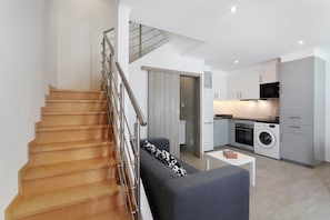 A modern apartment with a wooden staircase, a grey sofa, a kitchen with appliances, and a small table.
