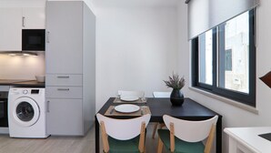 Une cuisine compacte équipée d’un lave-linge, d’une table à manger avec des chaises et d’une fenêtre donnant sur des immeubles.