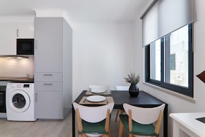 A compact kitchen with a washing machine, a dining table with chairs, and a window with a view of buildings.