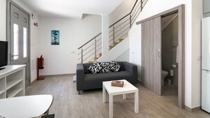 Un salon moderne avec un canapé gris, une table basse blanche, un escalier et une porte blanche donnant sur une salle de bain.
