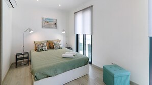 Une chambre à coucher comprenant un lit, des lampes de chevet, une fenêtre avec des stores, un petit tabouret et un climatiseur fixé au mur.