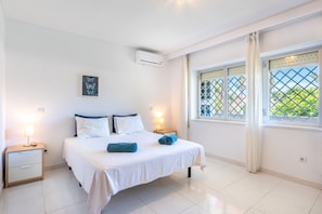 A bedroom with a bed, two bedside tables, a nightstand, a window with curtains, and an air conditioning unit.