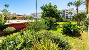 A landscaped garden with palm trees, a red building, and a multi-story white building in the background. - Apartment 'Eurosol Carihuela' with Shared Garden, Wi-Fi and Air Conditioning (Torremolinos)