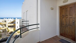 A balcony with a metal railing, a wooden door, and a view of other buildings and the sea.