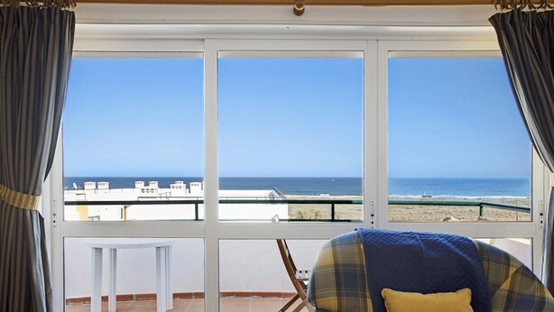 A room with a view of the sea, a chair with a blue and yellow pillow, and a small white table.