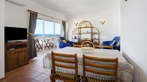 A room with a TV, dining table, chairs, a bookshelf, and a view of the sea.