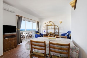 A room with a TV, dining table, chairs, a bookshelf, and a view of the sea.