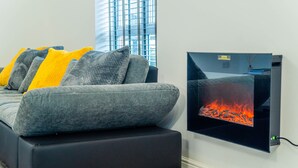 A modern living room with a grey sofa, yellow and grey cushions, and a wall-mounted fireplace with a visible fire. - TWO BEDROOM LUXURY GROUND FLOOR APARTMENT.  (London)