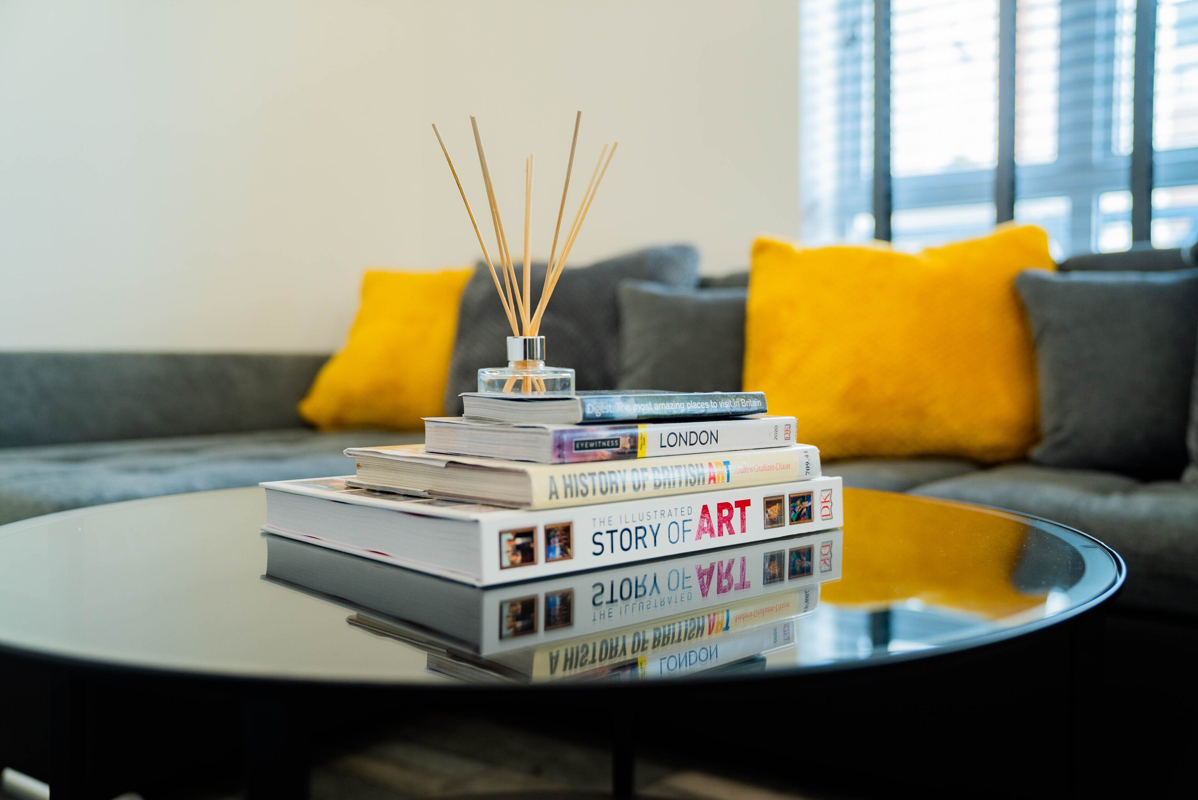 Ein Stapel Bücher auf einem Glastisch in einem modernen Interieur, inklusive eines grauen Sofas mit gelben Kissen.