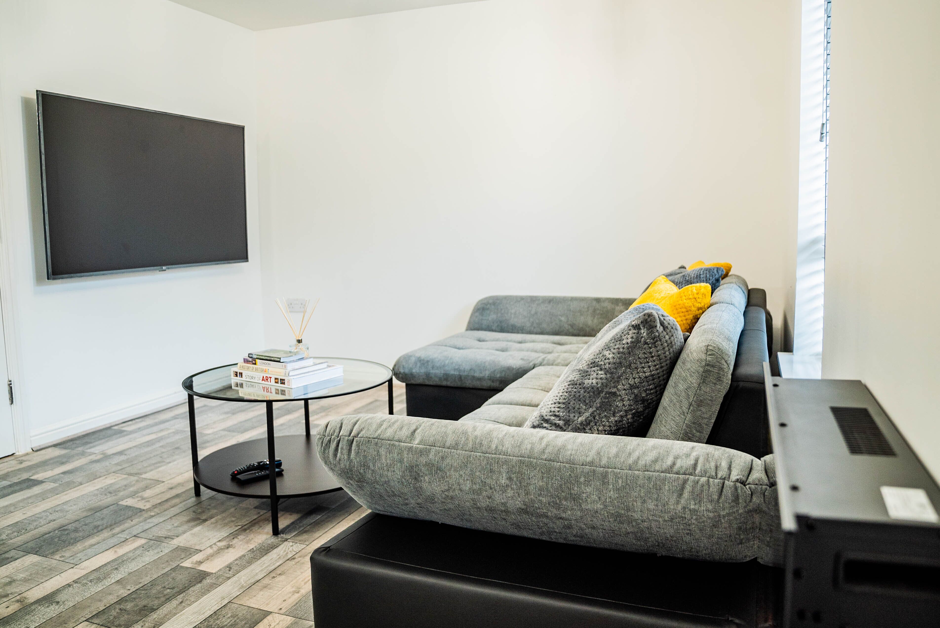 Een moderne woonkamer met een grijze bank, een flatscreen televisie, een klein rond salontafeltje en een boekenkast.