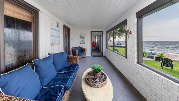 A covered outdoor area with a white ceiling, a wicker sofa with blue cushions, a wooden coffee table with a plant, and a view of the sea through large windows. - BAY HOUSE - Steps to its own Private Beach - Amazing Sunsets on the Water (Pensacola)