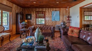 A wooden-paneled room with a leather sofa, a coffee table with books and decorative items, and a large window with a view of greenery. - BAY HOUSE - Steps to its own Private Beach - Amazing Sunsets on the Water (Pensacola)