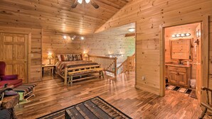A cozy cabin interior with a wooden floor, a bed with a colorful blanket, a red chair, a small table with a lamp, and a ceiling fan. - Dreamy Ellijay Resort Cabin w/ Game Room & Decks! (Ellijay)