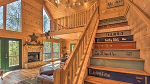 A wooden cabin interior with a fireplace, a staircase with books, and a view of the outdoors. - Dreamy Ellijay Resort Cabin w/ Game Room & Decks! (Ellijay)
