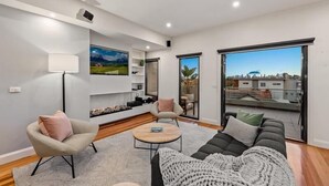 A modern living room with a sofa, armchair, and coffee table, a fireplace, and a city view through large windows. - StayCentral - Moonee Ponds Penthouse - Mount Alexander Rd, Moonee Ponds - 3 Bedrooms, 4 Beds, 2 Bathrooms, 2 secure undercover parking spot. (Moonee Ponds)