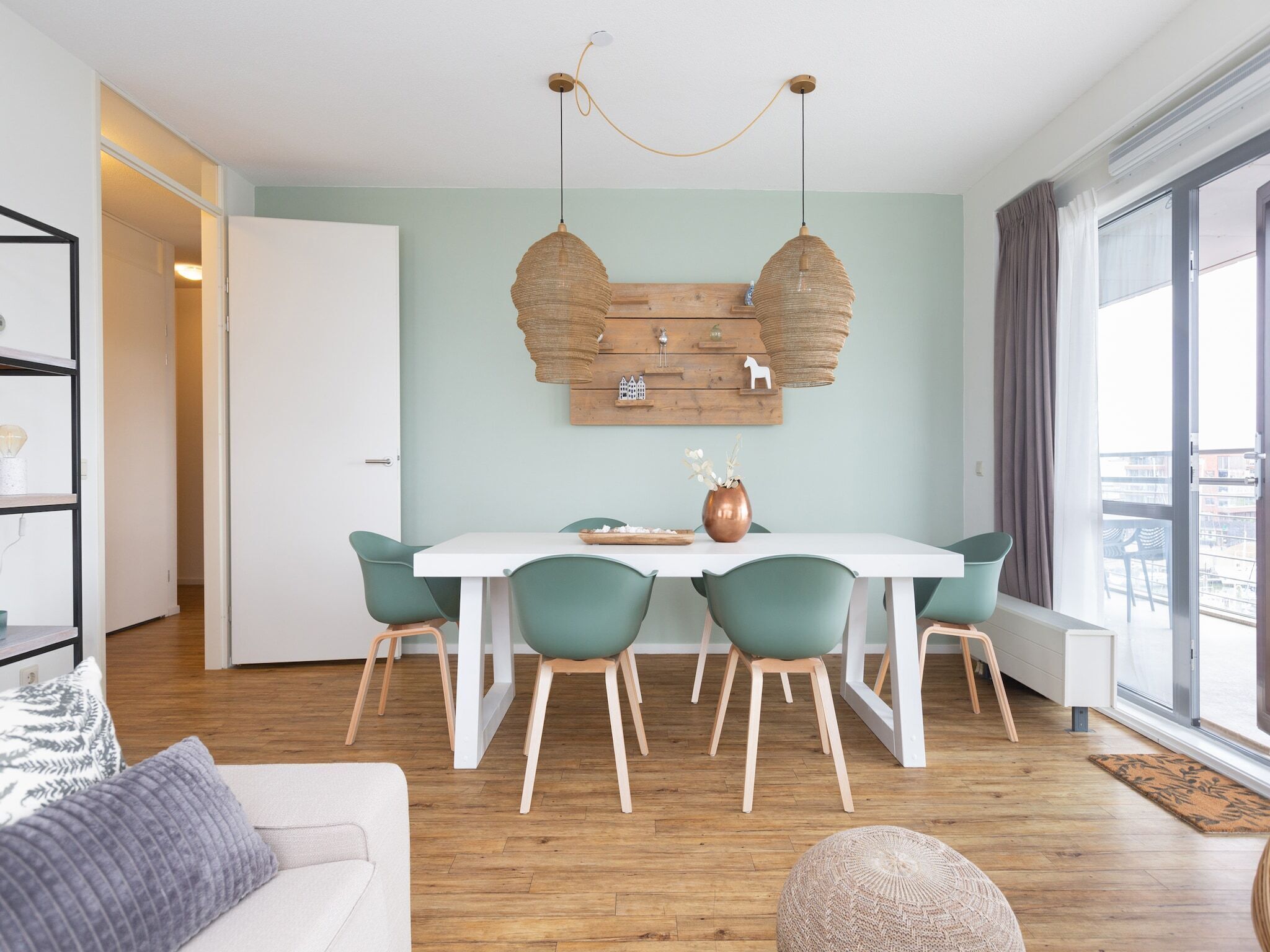 Un espace repas moderne avec une table blanche et des chaises turquoise, une œuvre d’art murale en bois au-dessus, et une grande fenêtre avec des rideaux.