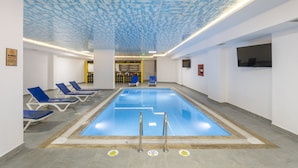 Une piscine intérieure à l’eau bleue, entourée de chaises longues et dotée d’un plafond décoré de motifs nuageux.