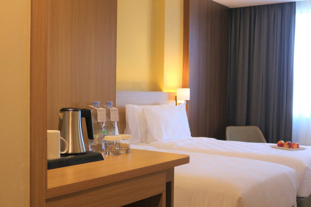 Ett hotellrum med två sängar, ett skrivbord i trä med en vattenkokare, glas och en mugg, en lampa och en fruktfat på nattduksbordet.