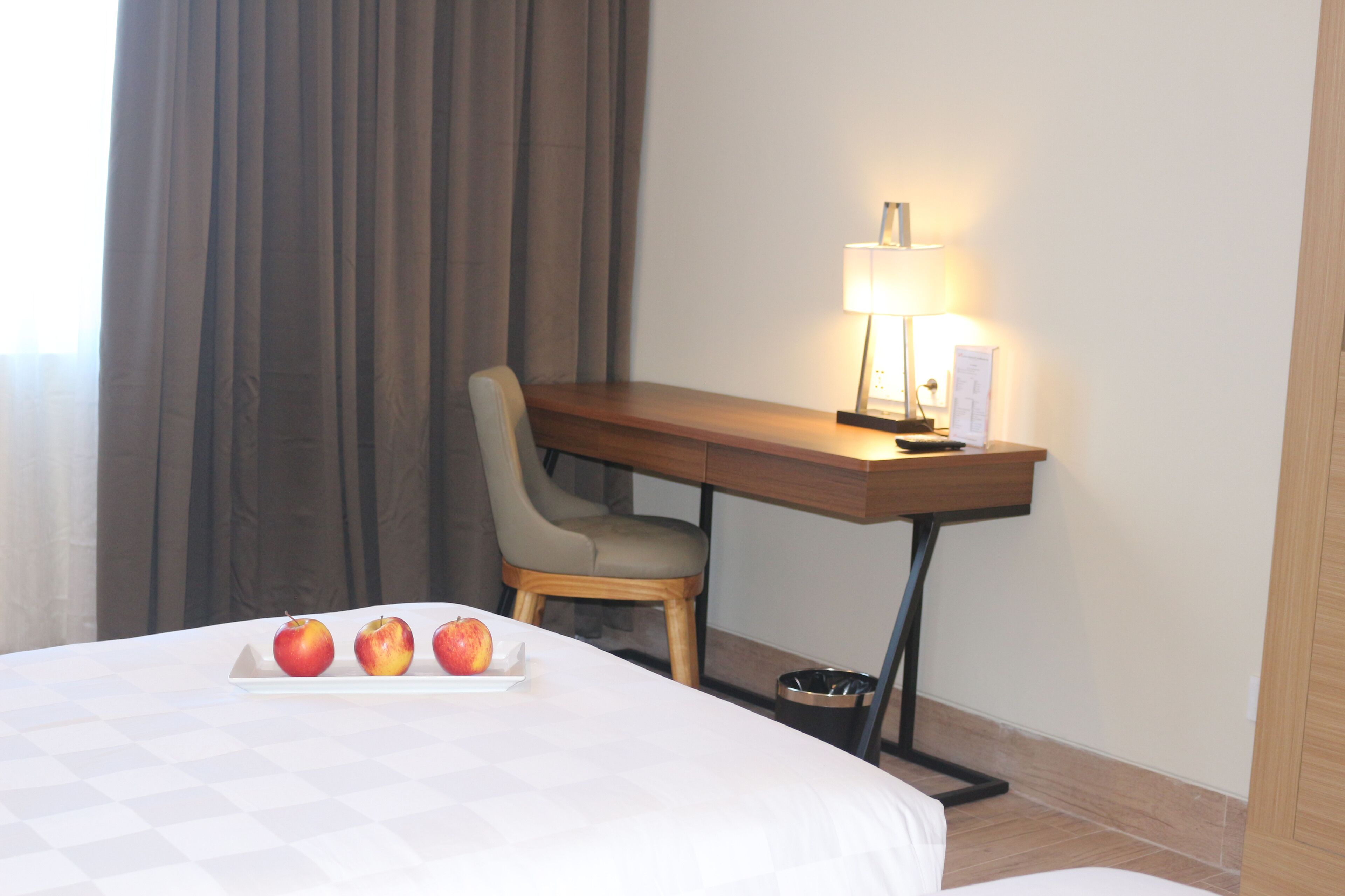 A hotel room with a bed, a desk with a lamp, a chair, and a tray with apples.
