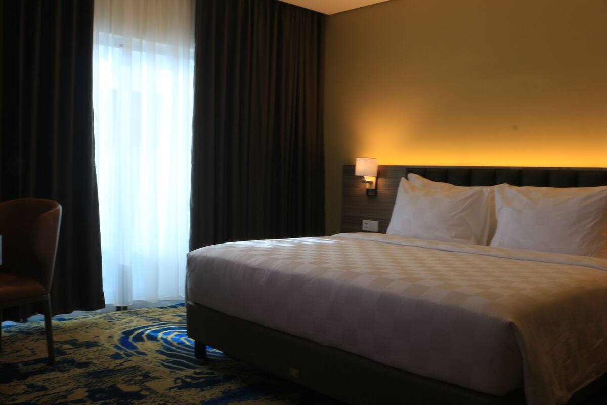 Ett hotellrum med en stor säng, en stol, en lampa och ett fönster med gardiner.