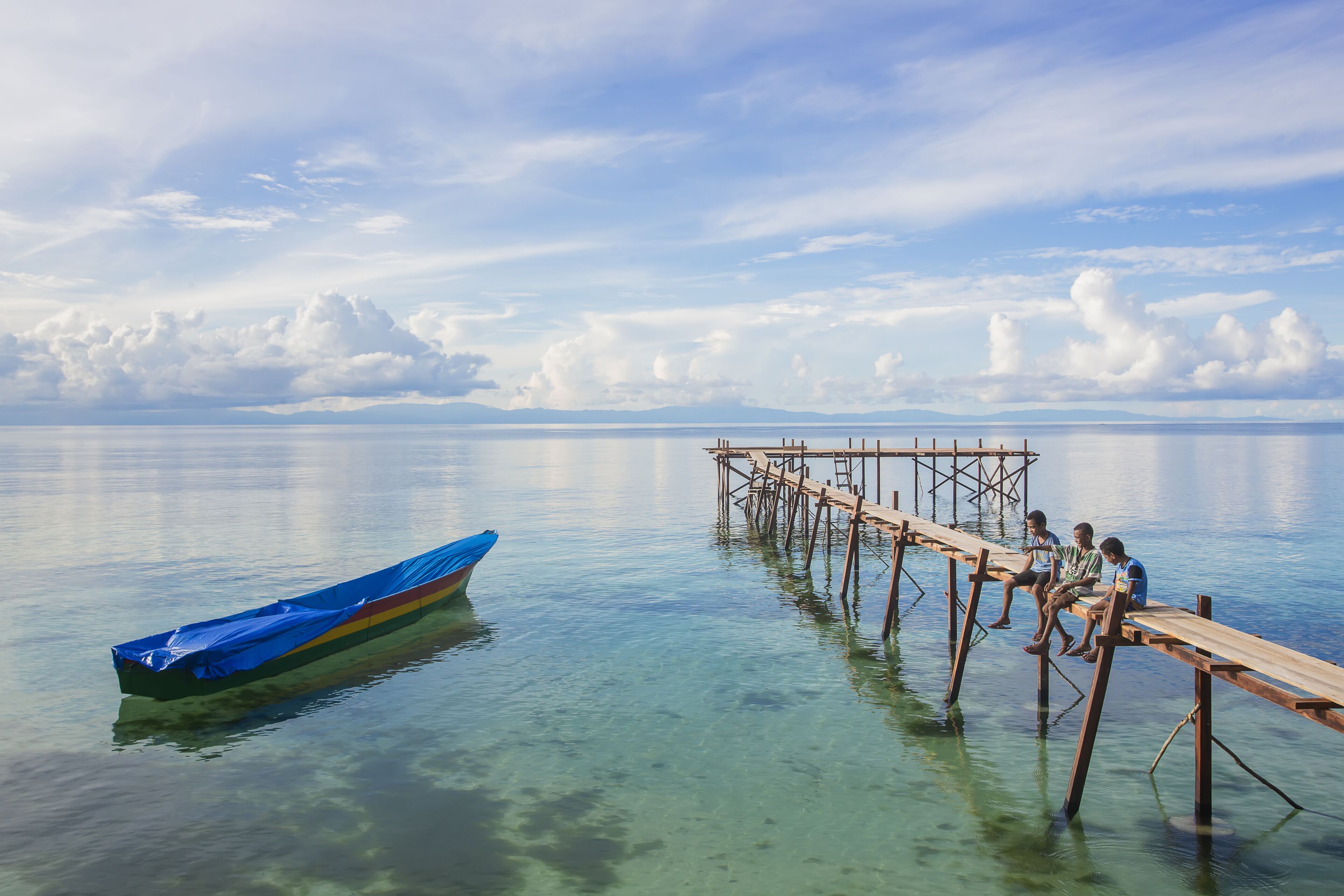 A wooden pier extending into calm water with a covered boat and people sitting on the pier.