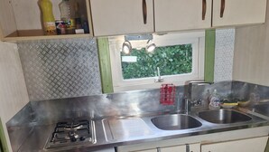 Cozinha compacta com fogão a gás, pia e pequena geladeira. Visível pela janela, há uma planta verde lá fora.