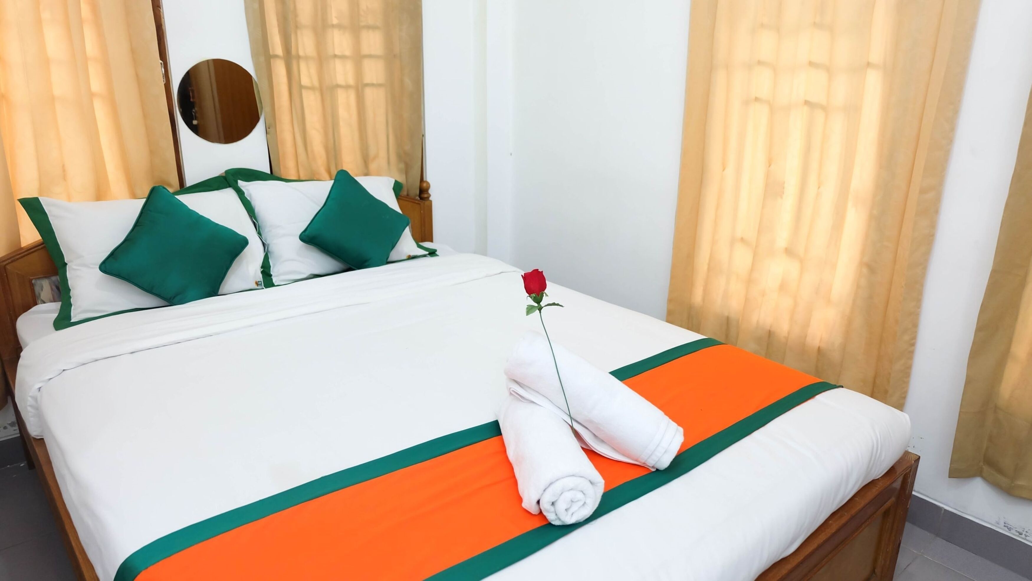 A neatly made bed with green and white bedding, a single rose, and rolled towels.