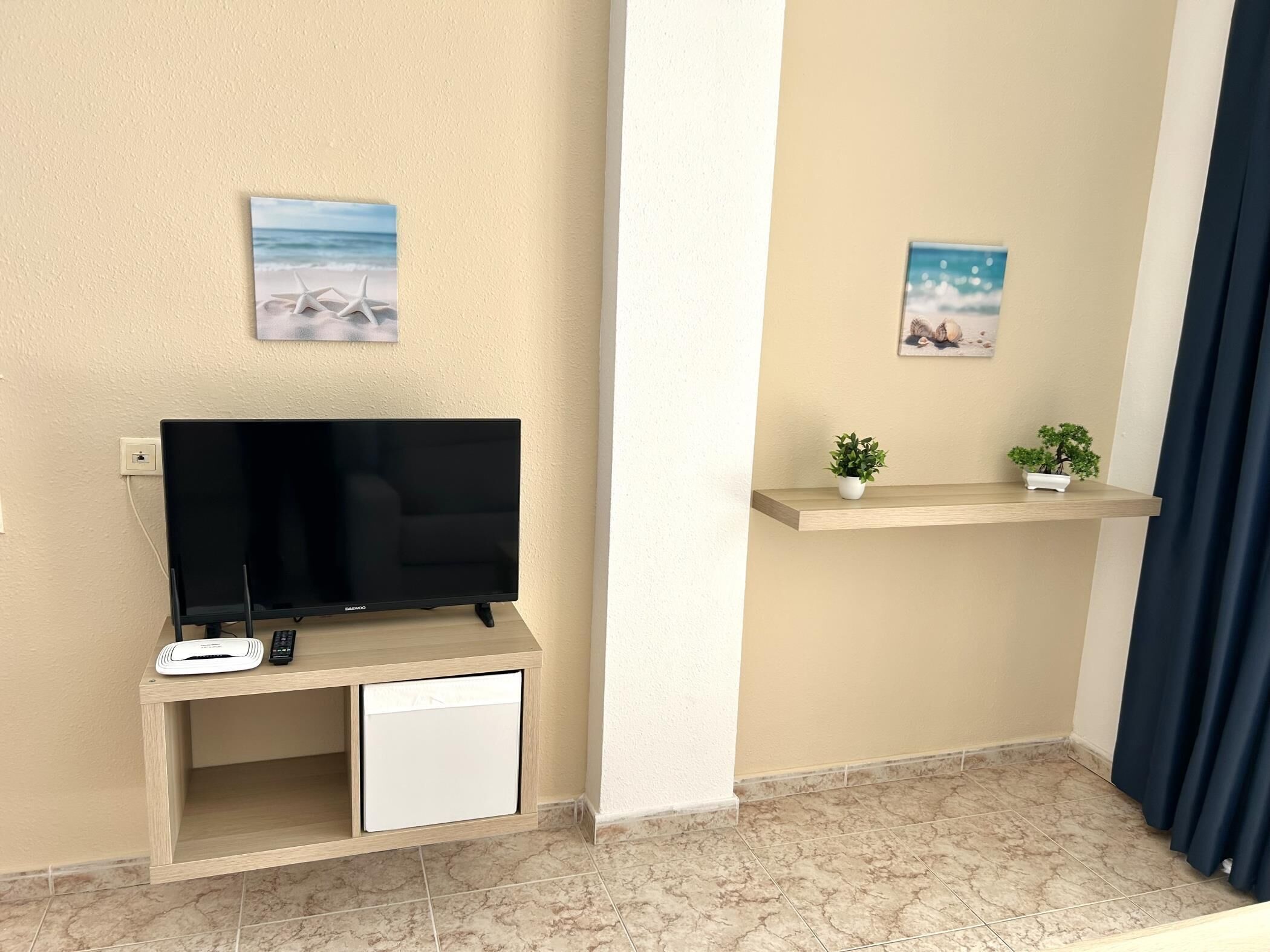 Un espace de vie comprenant un téléviseur à écran plat posé sur un meuble, un petit placard en dessous, et deux étagères murales ornées de plantes en pot.