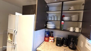 Eine kleine Küche mit einem weißen Kühlschrank, einer Kaffeemaschine, einem Toaster und einem Regal mit Geschirr und Gewürzen.