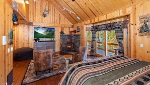 A cozy cabin interior with wooden walls, a fireplace, a television, and a large window with a view of the outdoors.