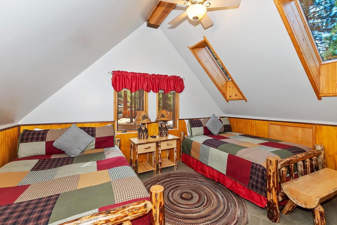 A cozy attic bedroom with two beds, a desk, and a window with a red valance.
